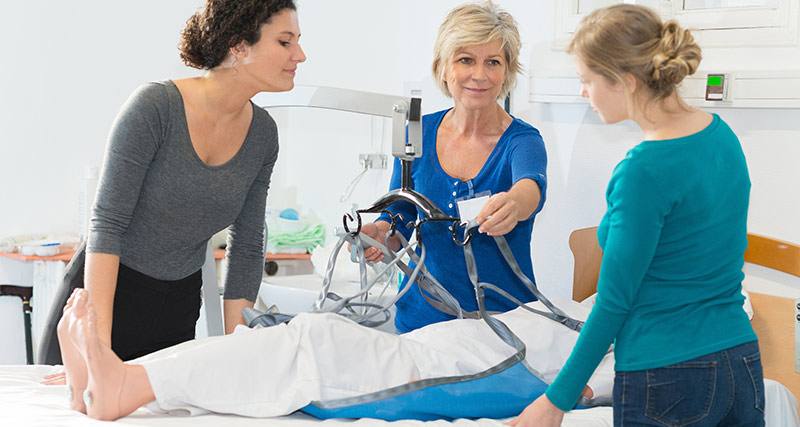 Zwei Pflegekräfte und eine Ausbilderin demonstrieren am Krankenbett den Einsatz eines Patientenlifters zum sicheren Umlagern.